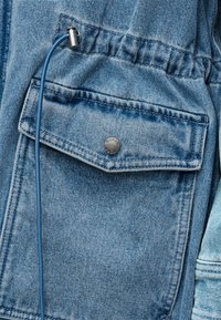 Μπουφάν ντένιμ με μπροστινή τσέπη που κλείνει με μεταλλικό κουμπί ασφαλείας, μέση με κορδόνι και λεπτομέρεια από μπλε κορδόνι. Ανοιχτό μπλε χρώμα με υφή υφάσματος.