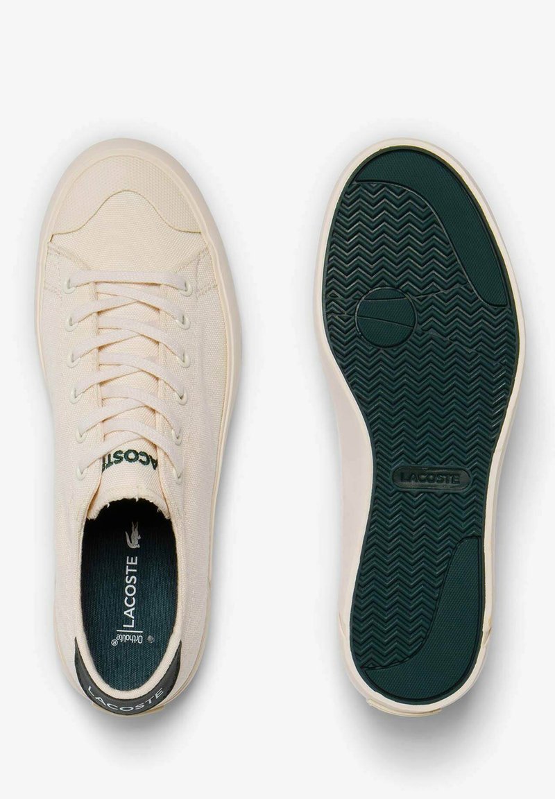 Sneakers in tela Lacoste di colore bianco sporco con punta tonda, dettagli in verde scuro a contrasto e suola in gomma nera testurizzata. Presenta sei fori per i lacci.