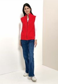 Rotes, gestricktes ärmelloses Weste mit einem Reißverschluss vorne, getragen über einem weißen Langarmshirt. Kombiniert mit blauen Schlagjeans und weißen Turnschuhen.