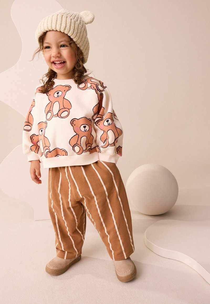 Light-colored sweatshirt with bear prints, brown striped pants, and beige shoes. Features a knitted cream beanie with a pom-pom top.