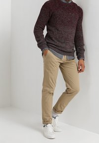 Pull en maille bordeaux foncé à finition texturée, associé à un pantalon chino beige clair et des baskets blanches. Tenue décontractée en superposition.