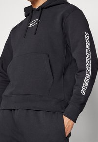 Svart sweatshirt med huva i bomullsblandning har en framficka, dragsko för huvan och vit "Nike Sportswear" text på ärmen.