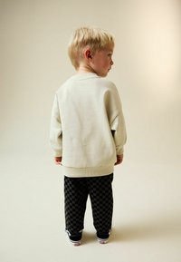 Bluză sport bej deschis cu manșete și tiv striate, combinată cu pantaloni în carouri negri și gri. Pantofi sport negri cu tălpi albe.