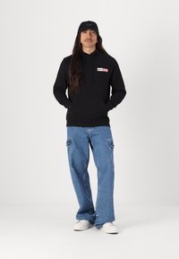 Sort hættejakke med logo, parret med lyseblå cargo jeans med store sidelommer. Båret med hvide sneakers og en sort kasket.