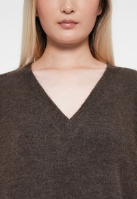 Bruine gebreide trui met een V-hals, zachte textuur en een ontspannen pasvorm. Kenmerken zijn een geribbelde halslijn en naden.