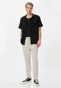 Camisa negra de manga corta con botones, superpuesta a una camiseta sin mangas negra, combinada con pantalones beige claros y zapatillas negras con suelas blancas.