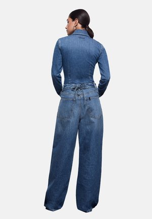 Donna con capelli scuri raccolti in una coda bassa, che indossa una giacca di jeans aderente e pantaloni di jeans a vita alta e ampi con dettagli allacciati sul retro.