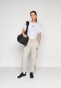 Hvit Adidas t-skjorte med logo, beige sweatpants med sideaksenter og svarte joggesko. Holder en svart duffelbag med skulderrem.