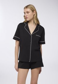 Completo pigiama corto nero con piping bianco, composto da una camicia con bottoni e pantaloni corti abbinati, realizzato in tessuto morbido.