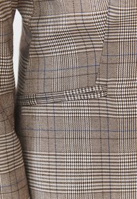 Hnědo-krémová kostkovaná látka s houndstooth a kostkovými vzory; texturovaný povrch s detailem propracované kapsy na boku.