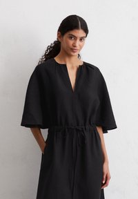 Abito in lino nero con maniche corte e larghe, scollo a V e vita con cordoncino. Presenta una vestibilità rilassata e tasche sui lati.