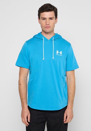 Młody mężczyzna noszący jasnoniebieską bluzę z kapturem z krótkim rękawem, białymi sznurkami i białym logo Under Armour na piersi.