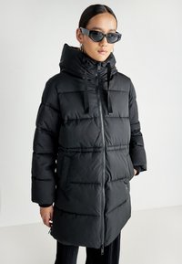 Svart polstret jakke med glidelås foran, justerbar hette og sidelommer. Har et quiltet design og kontrasterende svarte snorer.