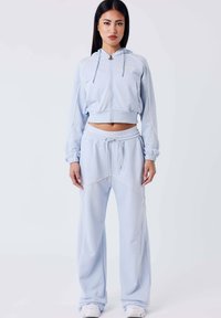 Vrouw staat, draagt een lichtblauwe zip-up cropped hoodie en een bijpassende wijdvallende sweatpants met witte sneakers, neutrale achtergrond.
