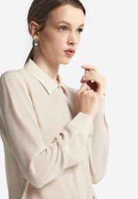 Beige, genomskinlig blus med klassisk krage, knäppning och långa ärmar. Har minimalistiska accenter i silverton vid ärmsluten och kragen.