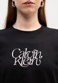 T-shirt en coton noir avec un logo stylisé blanc "Calvin Klein" sur le devant. Encolure ronde et manches courtes. Texture lisse.