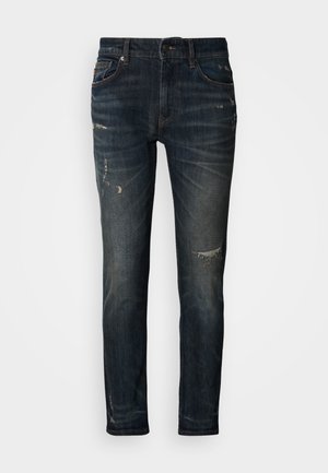 Tamno plave traperice uskog kroja, s istrošenim dijelovima i suptilnim blijeđenjem. Klasični dizajn s pet džepova, patentnim zatvaračem i kopčanjem na gumb.