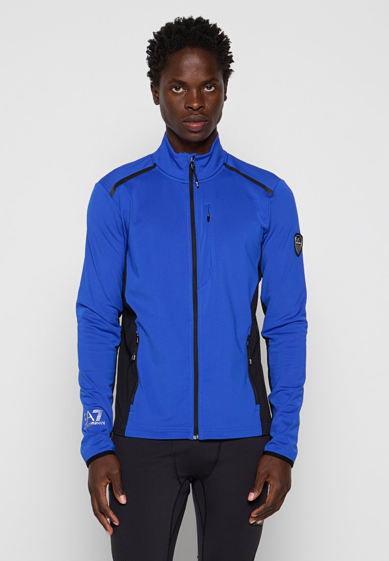Giacca softshell blu royal con accenti neri. Presenta una chiusura frontale con zip, due tasche laterali e una tasca piccola sul petto. Tessuto elasticizzato.
