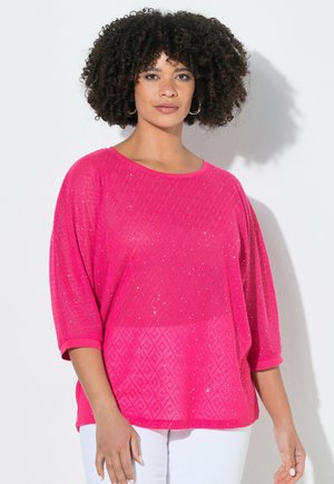 Kobieta o kręconych włosach, nosząca kolczyki-koła i jaskraworóżową, fakturowaną bluzkę z rękawem do połowy ramienia oraz białe spodnie, stoi na białym tle.