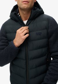 Gilet imbottito blu navy con cappuccio, parte superiore imbottita, maniche in maglia testurizzata, chiusura con zip e logo "SUPERDRY" sul petto.