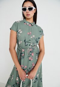 Robe florale en tissu vert pâle avec des motifs de fleurs roses et bleues ; manches courtes ; taille ceinturée ; associée à des lunettes de soleil blanches surdimensionnées.