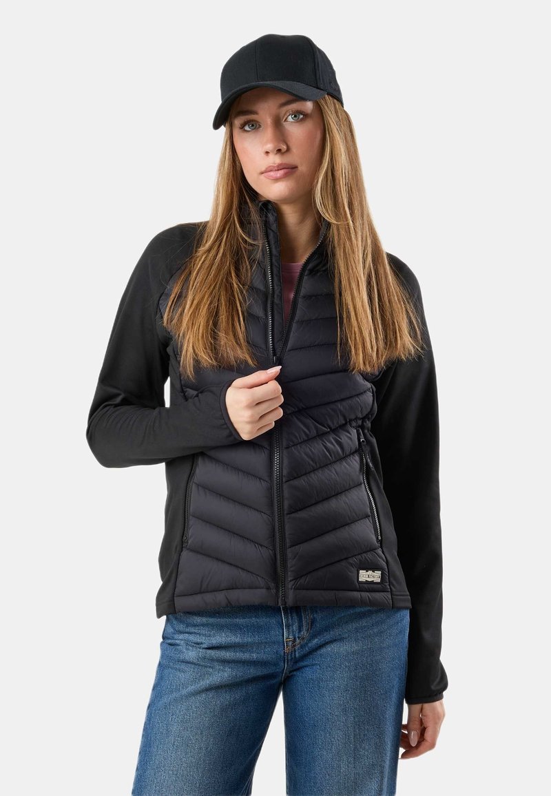 Jeune femme portant une casquette noire, une veste matelassée noire avec fermeture éclair et un jean bleu, debout devant un fond uni.