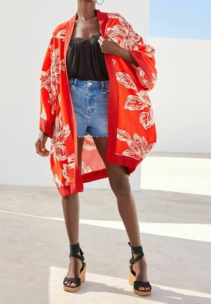 Červené květované kimono s bílými vzory, nošené přes černý top a džínové šortky. Má široké rukávy a složitou lemování. Nosit s černými sandály na klínu.
