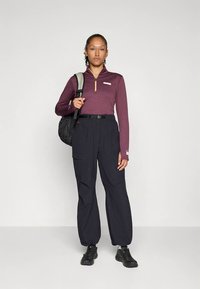 Donna in piedi che indossa una maglietta a maniche lunghe borgogna con zip, pantaloni cargo neri, sneakers nere e porta uno zaino nero su una spalla.