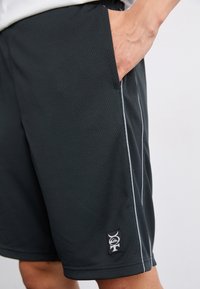 Sorte sportshorts laget av teksturert stoff, med sidelommer, en hvit aksentstripe og et lite logopatch nær nederkanten.