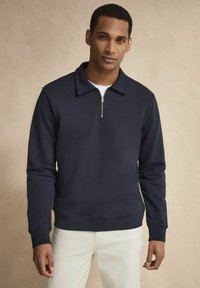Mörkblå tröja med zip-krage, tillverkad av en mjuk tyg. Har ribbade muddar och nederkant, samt en avslappnad passform, bärs över en vit skjorta.