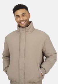 Giacca puffer beige con colletto alto, chiusura frontale con zip, polsini elasticizzati e tasche laterali. Materiale testurizzato e isolato. Vestibilità casual.