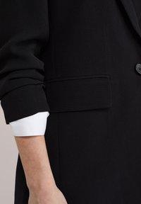 Černý blazer se strukturovaným designem, s bílým manžetovým lemem vykukujícím z rukávu a jedním černým knoflíkem jako akcentovým prvkem.
