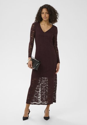 Moteris dėvi ilgomis rankovėmis tamsiai violetinę nėriniuotą suknelę su V formos iškirpte, laiko juodą delninę ir dėvi juodus smailėjančius aukštakulnius.