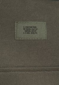 Étiquette en tissu verte cousue sur un textile tricoté vert olive, comportant un texte imprimé en noir avec les détails de la marque et du produit.
