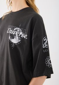 T-shirt preta de manga curta com estampado gráfico branco da lua, texto "UNREAL Universe" e logótipos nos mangas. Material de algodão.