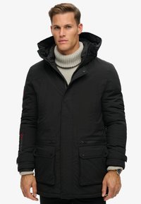 Chaqueta tipo plumas negra con capucha desmontable, cierre frontal y múltiples bolsillos. Presenta un cuello de punto acanalado y un acento de logo rojo en la manga.