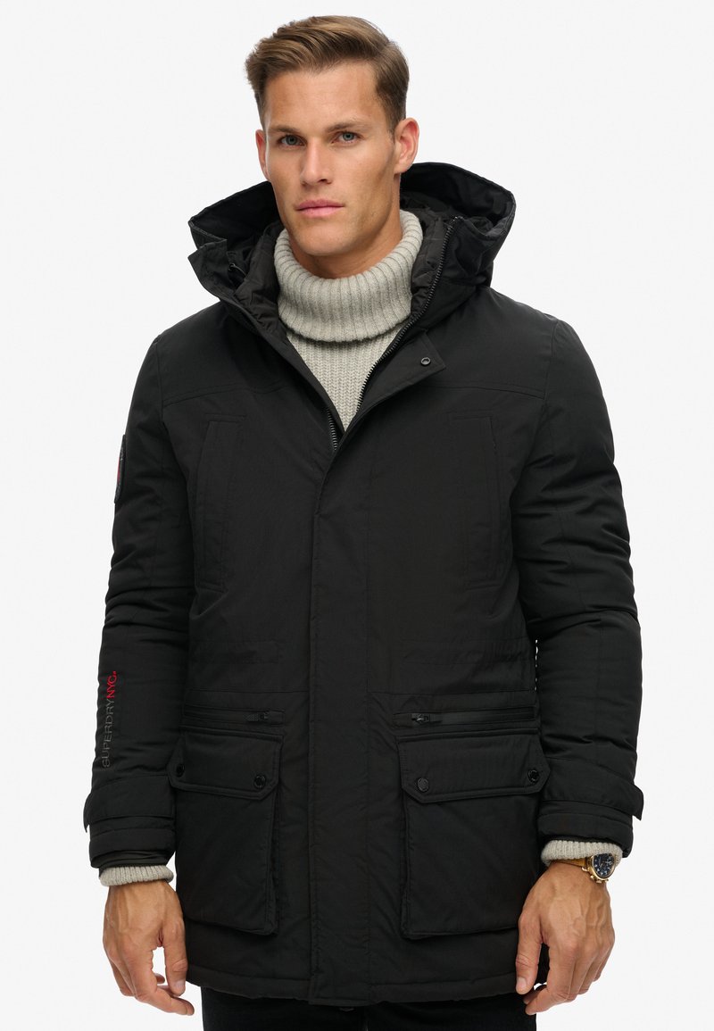 Chaqueta tipo plumas negra con capucha desmontable, cierre frontal y múltiples bolsillos. Presenta un cuello de punto acanalado y un acento de logo rojo en la manga.