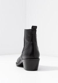Czarne skórzane botki za kostkę z niskim, grubym obcasem, pętelką z tyłu i zapięciem na zamek, wyeksponowane na białej powierzchni.