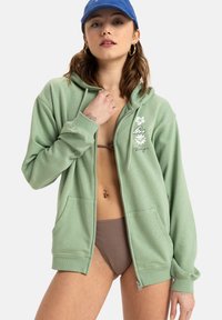 Ljusgrön zip-up huvtröja med en framficka, prydd med vita blommor och vågmönster. Mjuk tygkvalitet och avslappnad passform.
