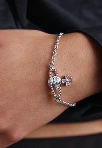 Pulseira de corrente prateada com um pendente redondo com gemas rosa e um pequeno encantamento de cruz. Elos texturizados e um acabamento polido.