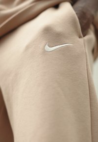 Pantalones de chándal Nike en tejido beige claro, con una textura suave y un logotipo bordado en blanco en el muslo.