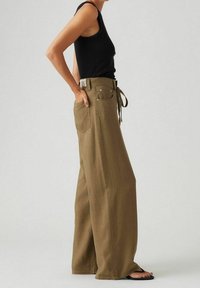 Pantalones de pierna ancha en tela verde oliva, que cuentan con una cintura alta, cinturón de lazo y detalles de bolsillos sutiles. Combinados con una blusa negra ajustada sin mangas.