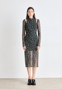 Green eye print midi dress with a high neck, long sheer sleeves, and side slits. Made of lightweight, transparent fabric. Black shoes worn.