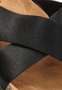 Sandalias de cuero negro con tiras entrelazadas de textura mate, apoyadas sobre una suela marrón con textura.