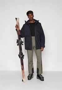 Skier in a dark navy jacket with large pockets, olive green pants, and black ski boots. Holding a light ski with black bindings.