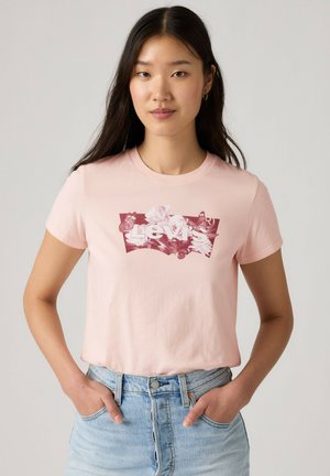Ung kvinde med langt mørkt hår iført en lyserød Levi's t-shirt med blomsterprint-logo og lyseblå højtaljede jeans med hænderne i lommerne.