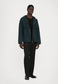 Άνδρας με σκοτεινή κουκούλα, μαύρο ριγέ πουλόβερ, μαύρα παντελόνια και μαύρα κοστούμια παπούτσια, όρθιος μπροστά σε λευκό φόντο.