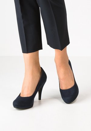Kvinde iført sorte cropped bukser og sorte semskede højhælede pumps, stående på et hvidt gulv.