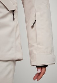 Beige vandtæt jakkeærme med sorte lynlåstræk, elastisk manschet og broderet logo i kanten. Glat tekstur, tætsiddende design.