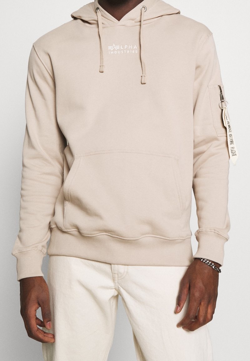 Personne portant un sweat à capuche beige Alpha Industries avec une poche avant et une poche zippée sur la manche avec une étiquette "Remove Before Flight".